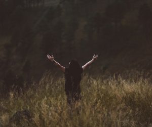 lady standing in a field facing a forest with arms upstreached to the sky